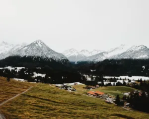 Landscape with a few houses in the foreground in front of a snow-covered mountain range. The snow line begins exactly halfway up the image.
